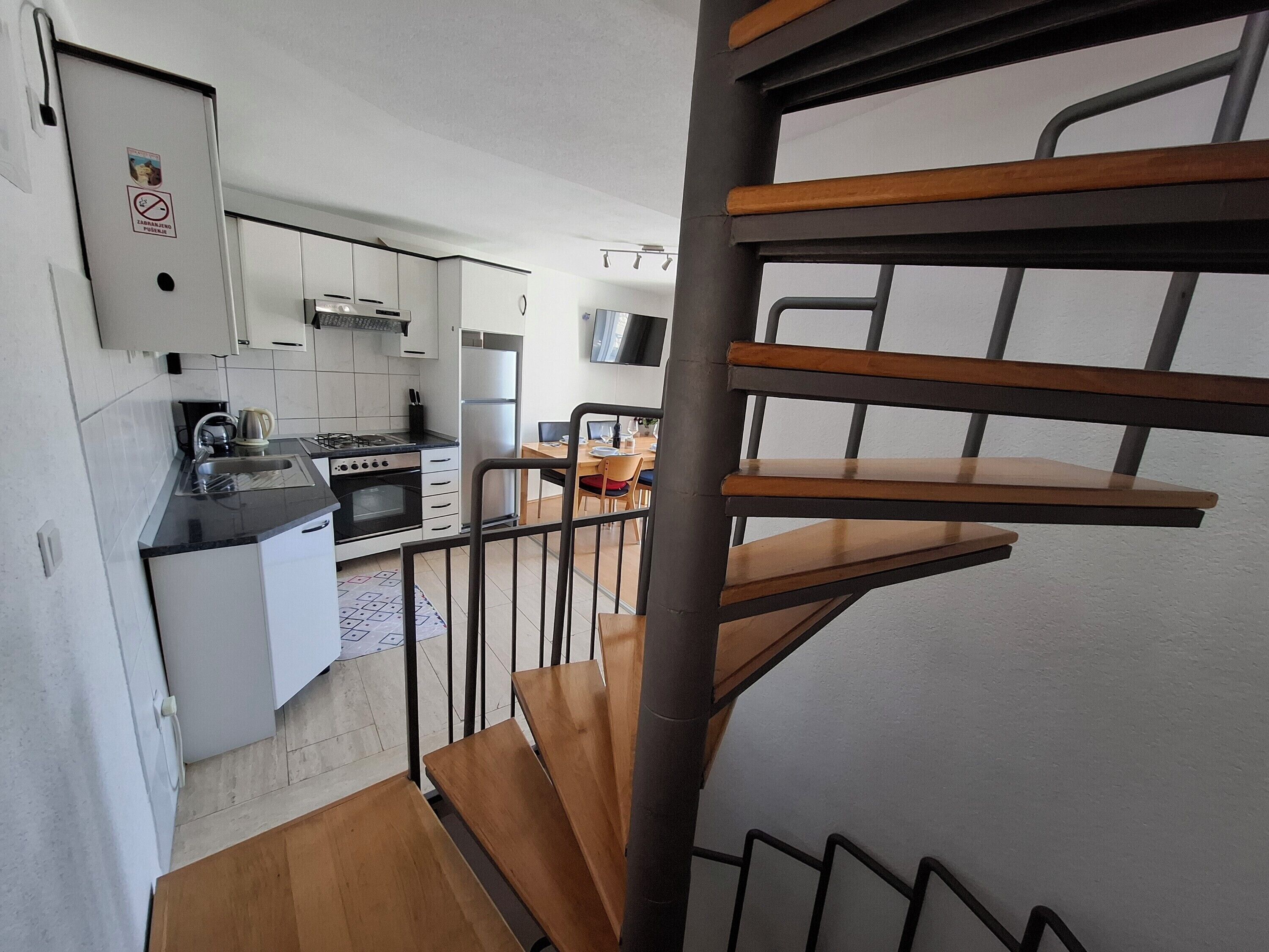 A modern kitchen with white cabinets, a stove, and a sink. There is a dining area with a table and chairs. A wooden staircase leads to an upper level.