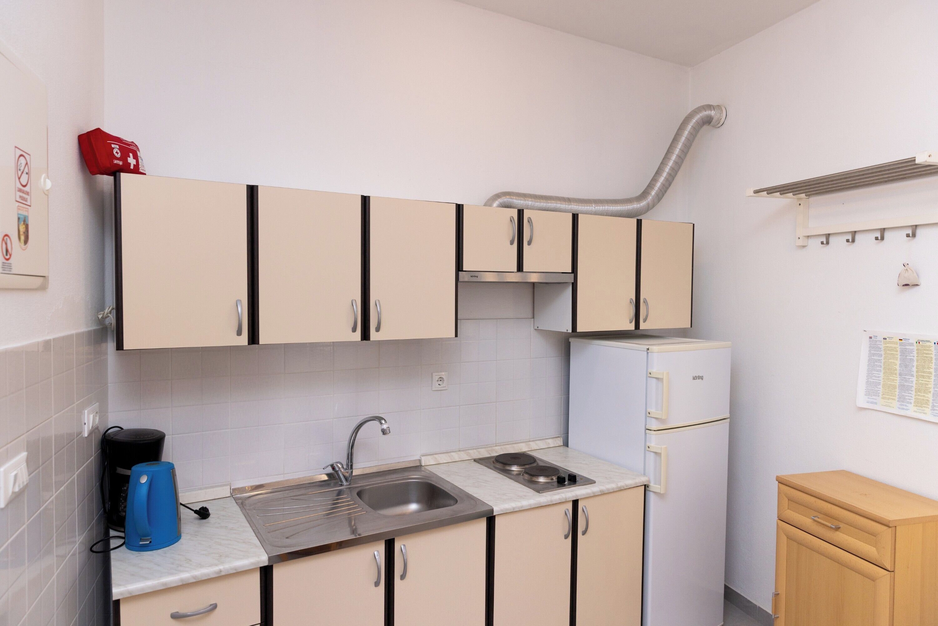Una cucina compatta con mobili beige, un lavello in acciaio inossidabile, un piano cottura integrato e un piccolo frigorifero.