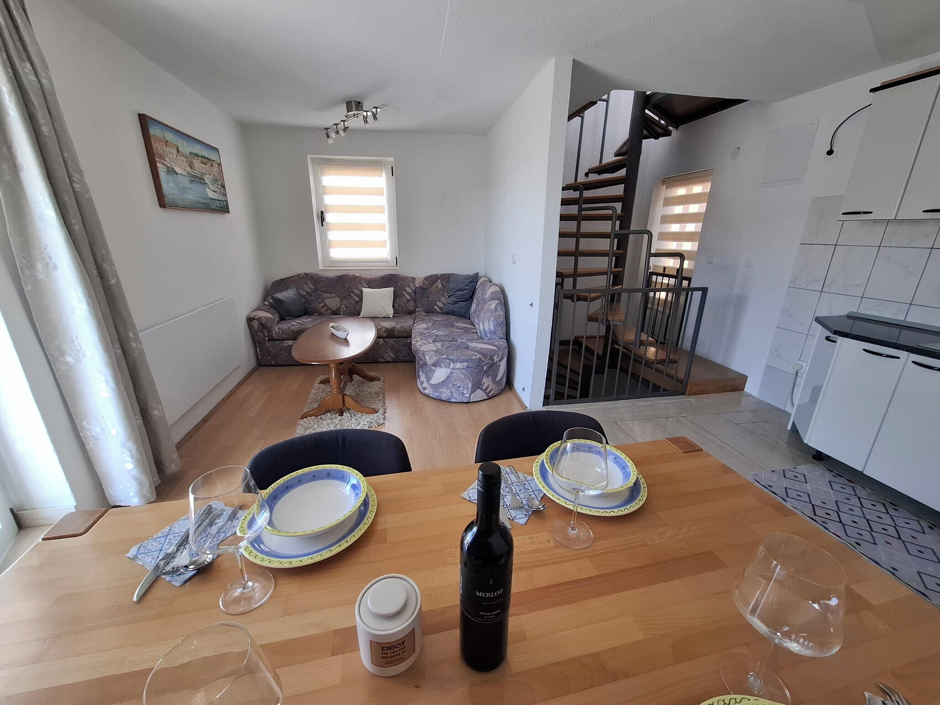 A dining area with a wooden table set for two, a bottle of Merlot wine, and a small white container.