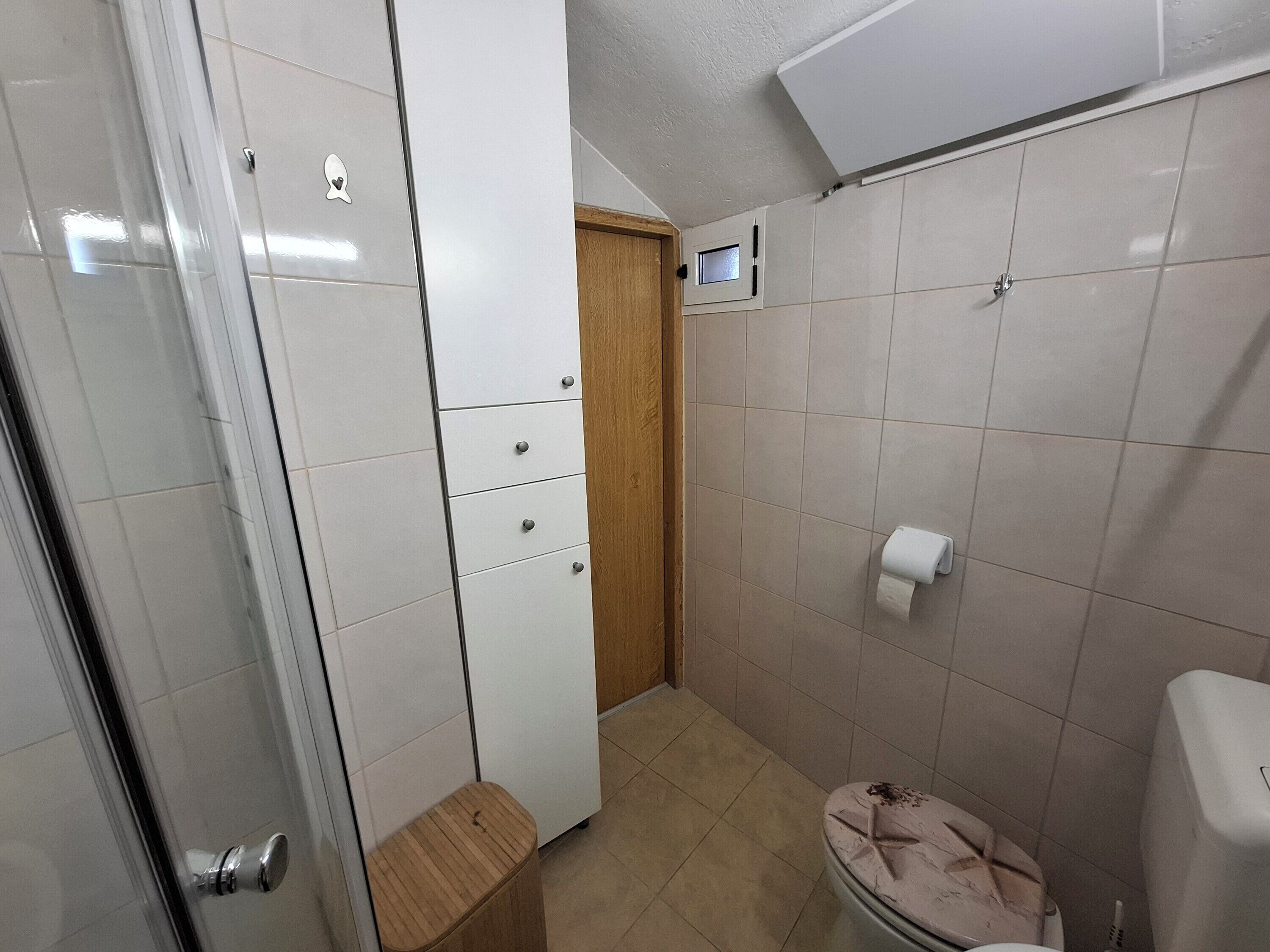 A bathroom with a toilet, a small wooden stool, a white cabinet with drawers, and a shower area.