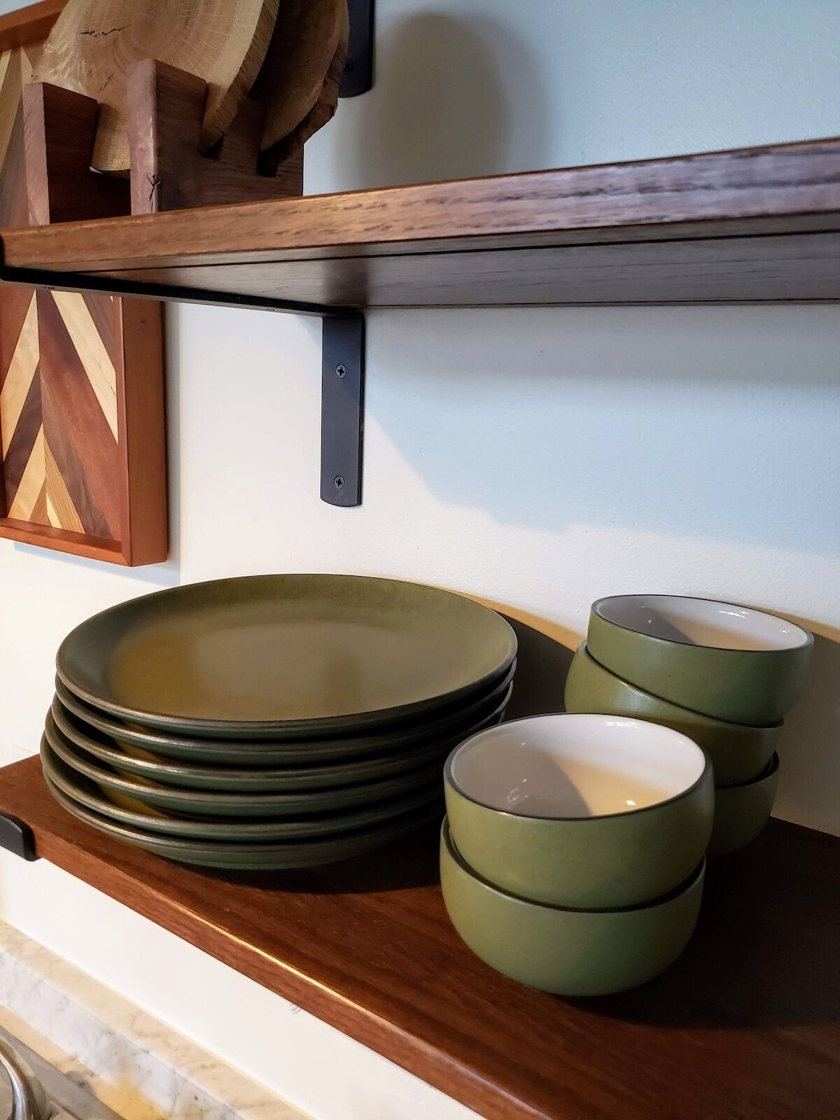 Un comptoir de cuisine avec des assiettes et des bols empilés, une étagère en bois au-dessus et un interrupteur fixé au mur.