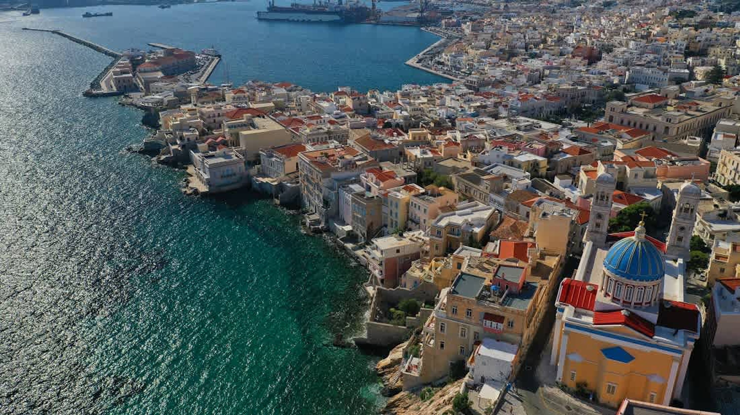 An aerial view of a coastal city with dense urban buildings, a prominent church with a blue dome, and a large ship docked in the harbor.