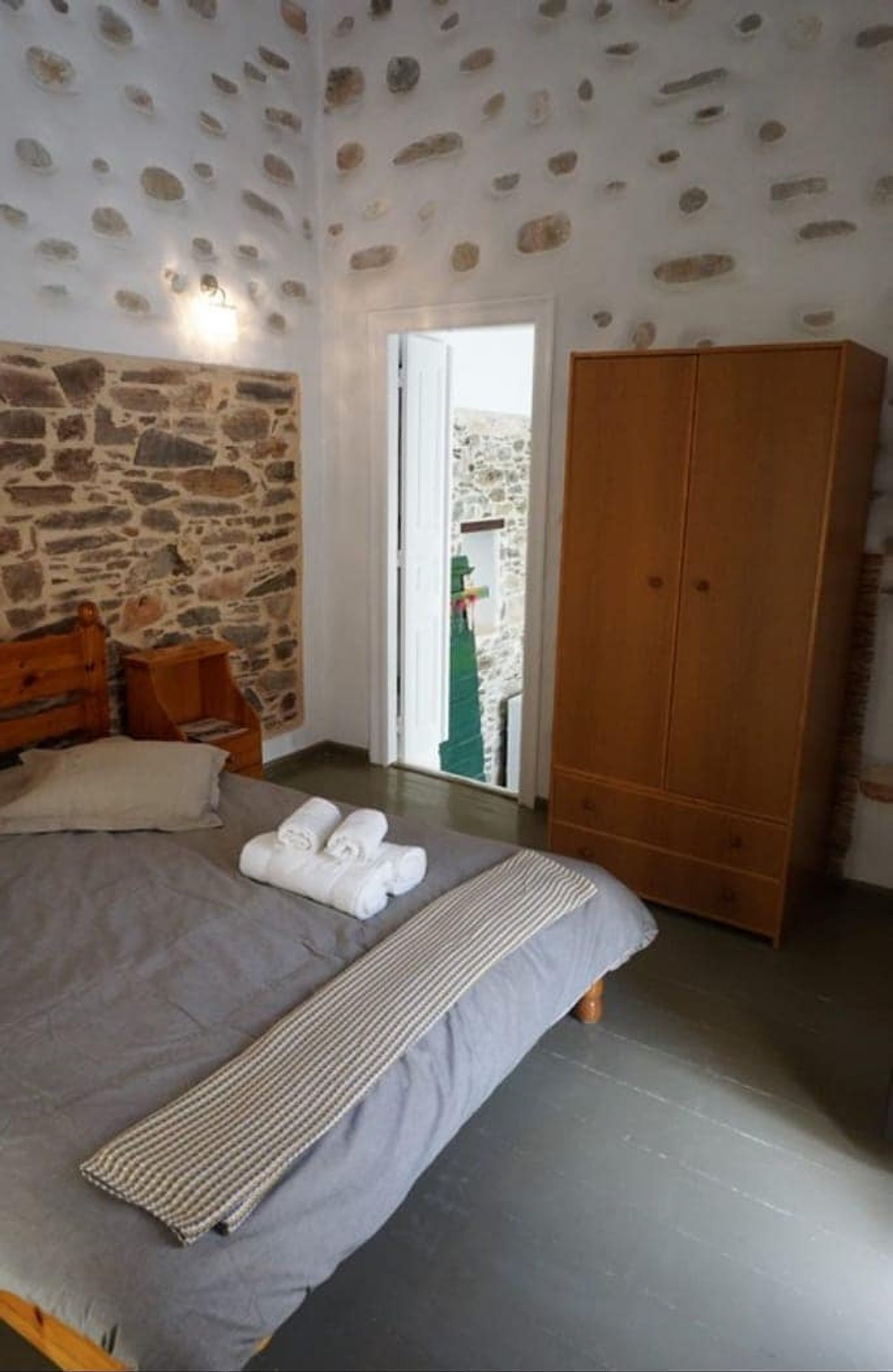 A bedroom with a stone wall, a wooden wardrobe, a bed with a grey bedspread, and a view of the outside.