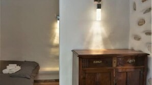 Una habitación con piso de piedra, un aparador de madera con cajones, una cama con manta gris y una lámpara fijada a la pared.