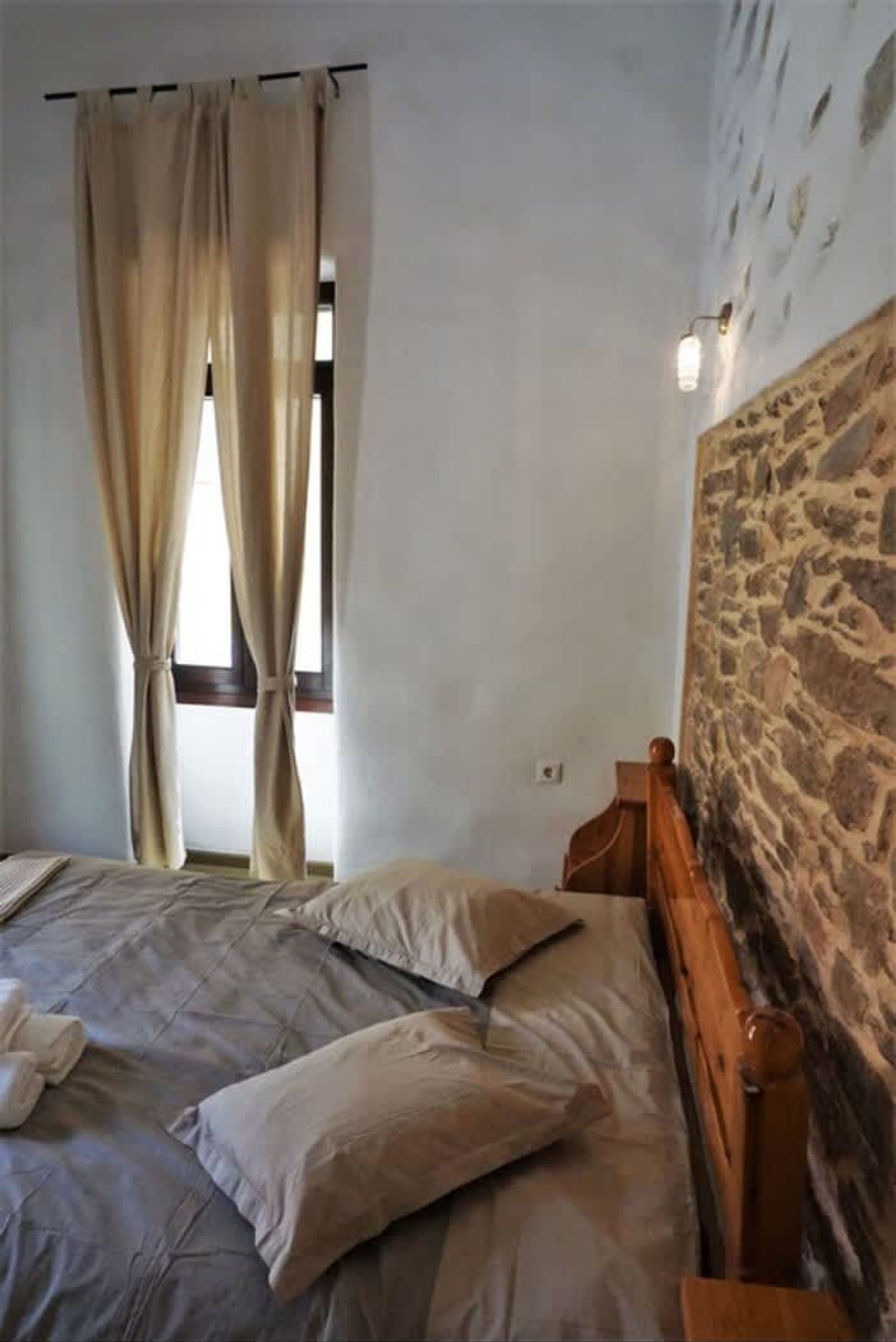 A bedroom with a bed, beige curtains, a stone wall, and a small window.