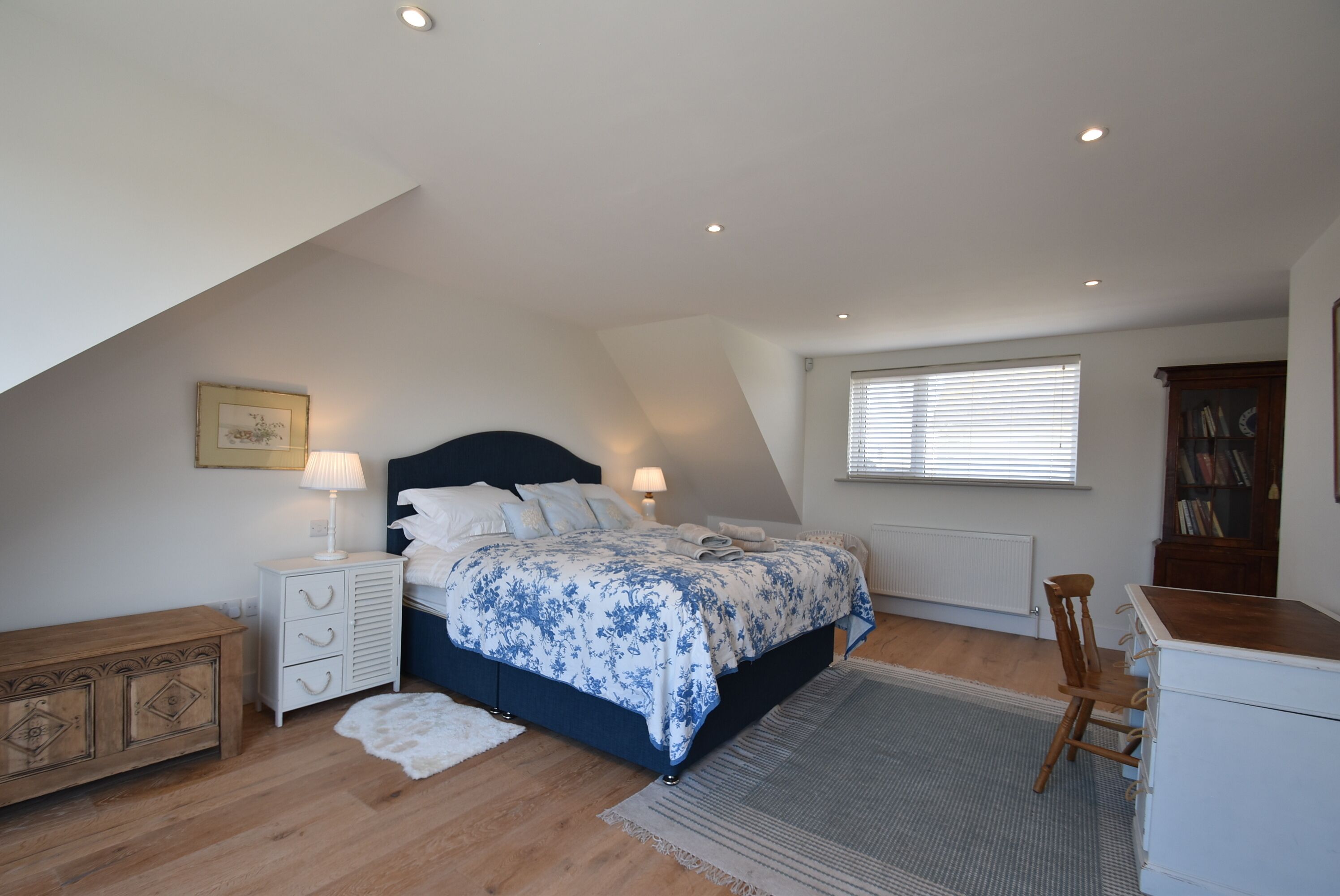 Un dormitorio con cama, mesitas de noche, cómoda de madera, radiador, ventana con persianas y estantería.