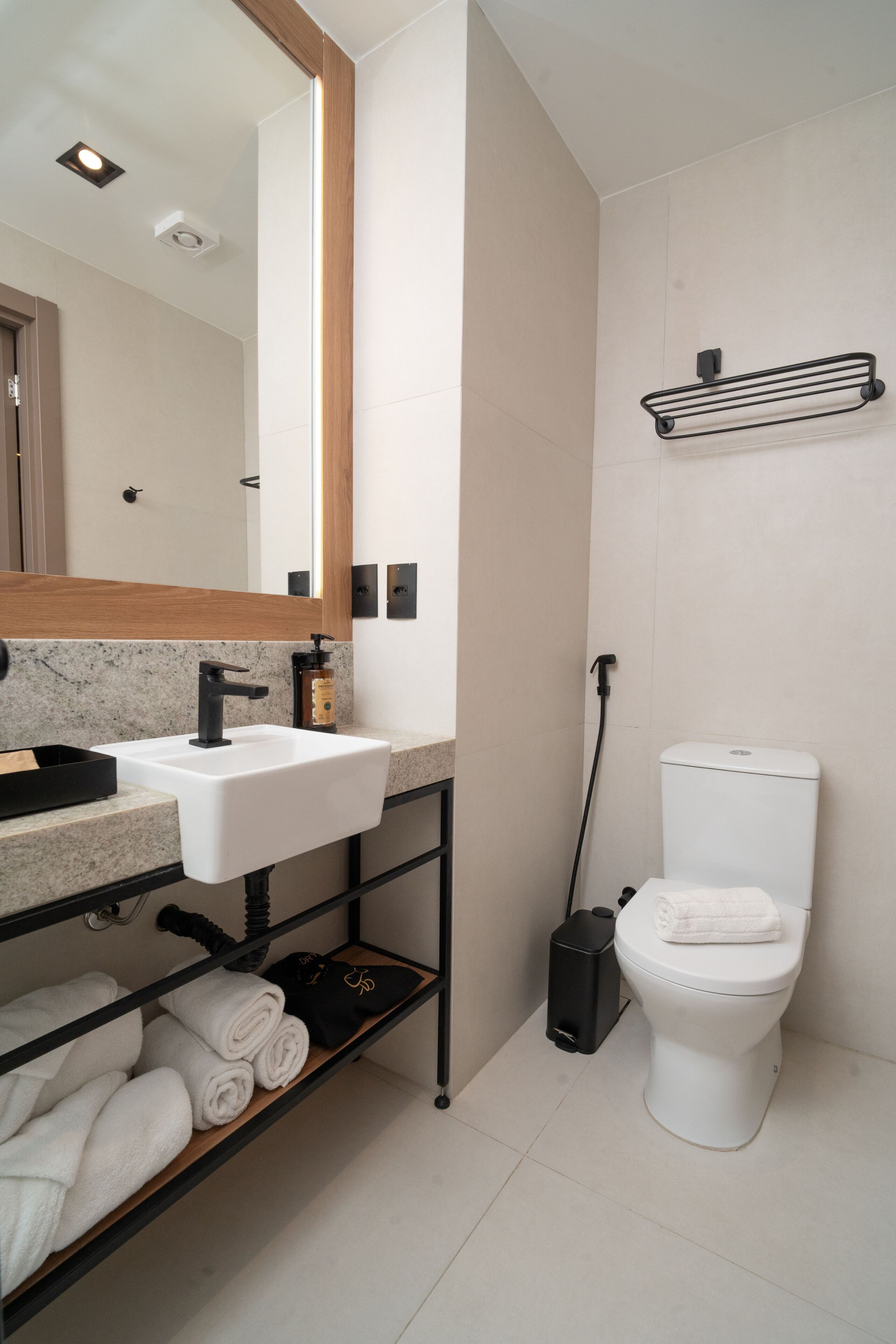 Et moderne badeværelse med en hvid vask, en sort hane og et toilet. Der er en hylde med håndklæder og en sort skraldespand.