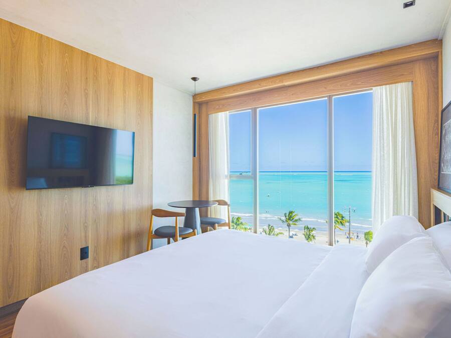 A hotel room with a large bed, a TV mounted on the wall, a small table with chairs, and a large window offering a view of the beach and ocean.