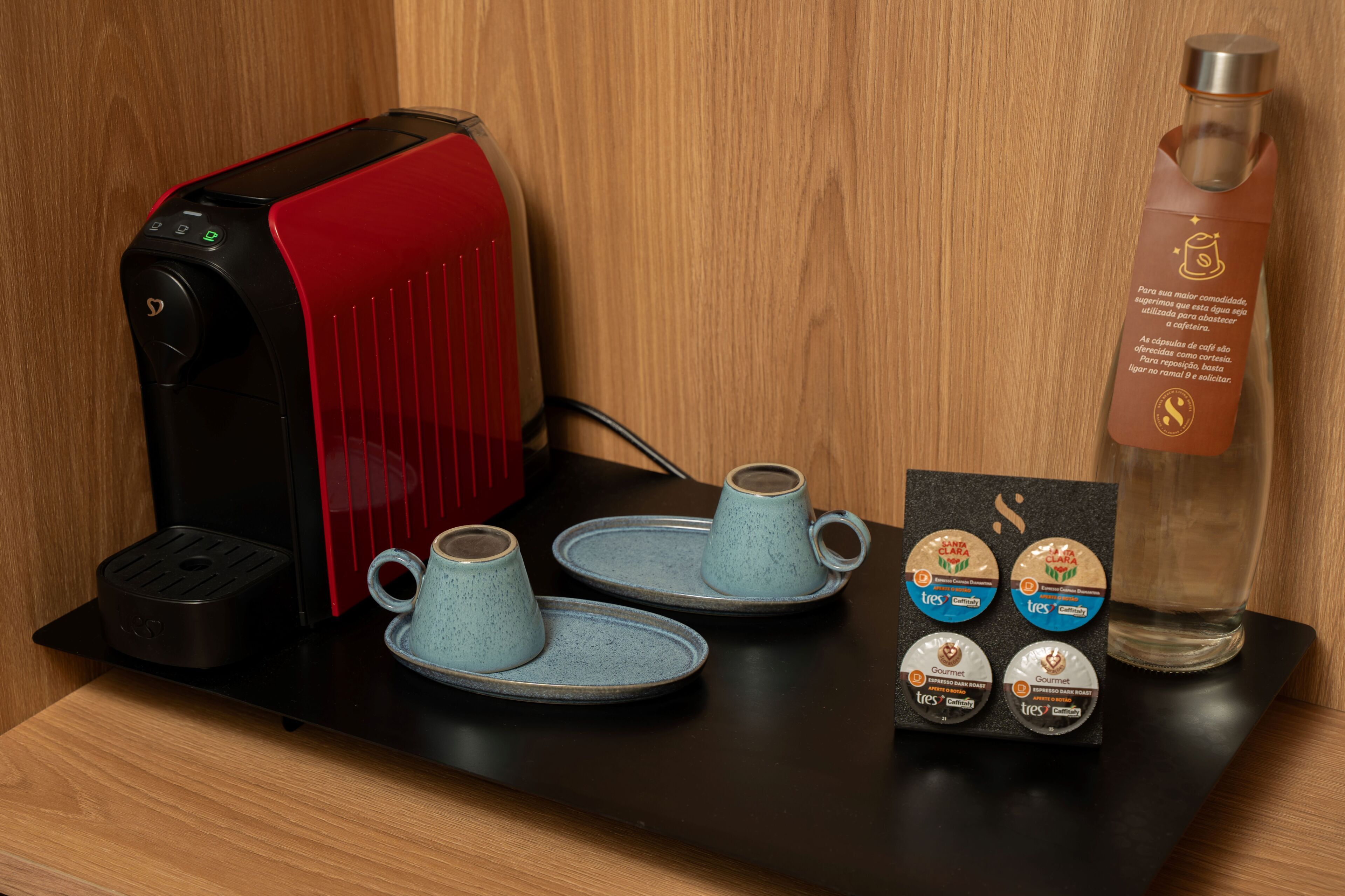 A coffee maker, two ceramic cups, a glass of water, and a card with coffee pods.