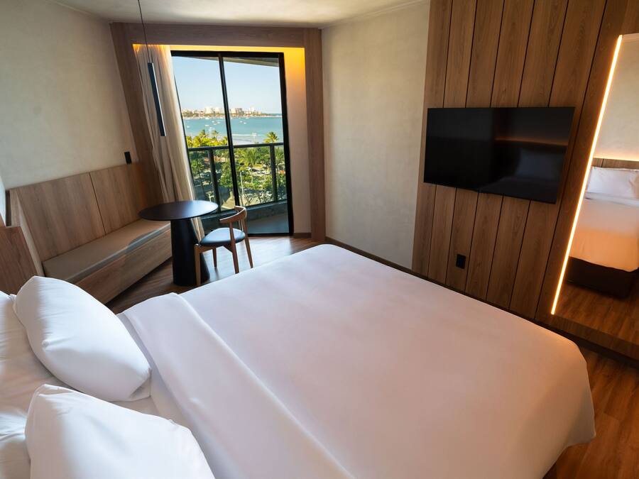 Habitación de hotel con una cama grande, un televisor montado en la pared, una mesita pequeña y un balcón con vistas.
