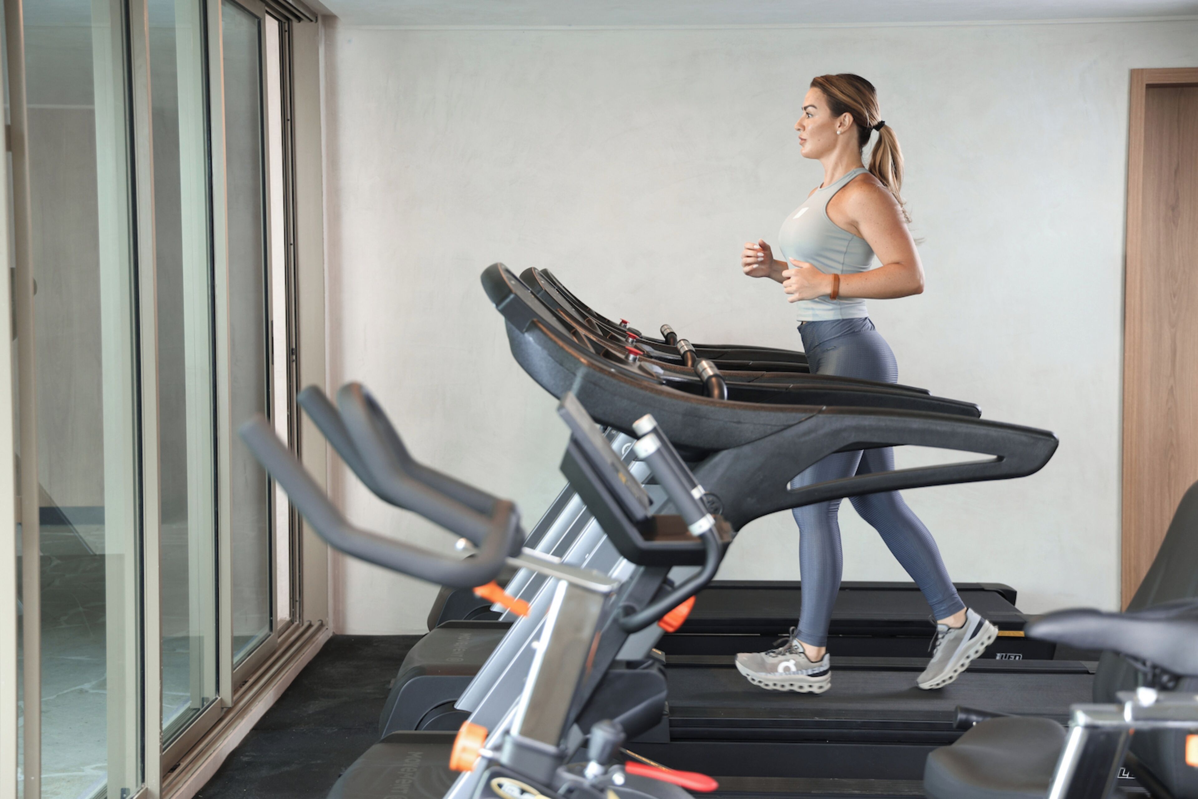 A woman running on a treadmill in a gym with multiple treadmills and a glass wall.