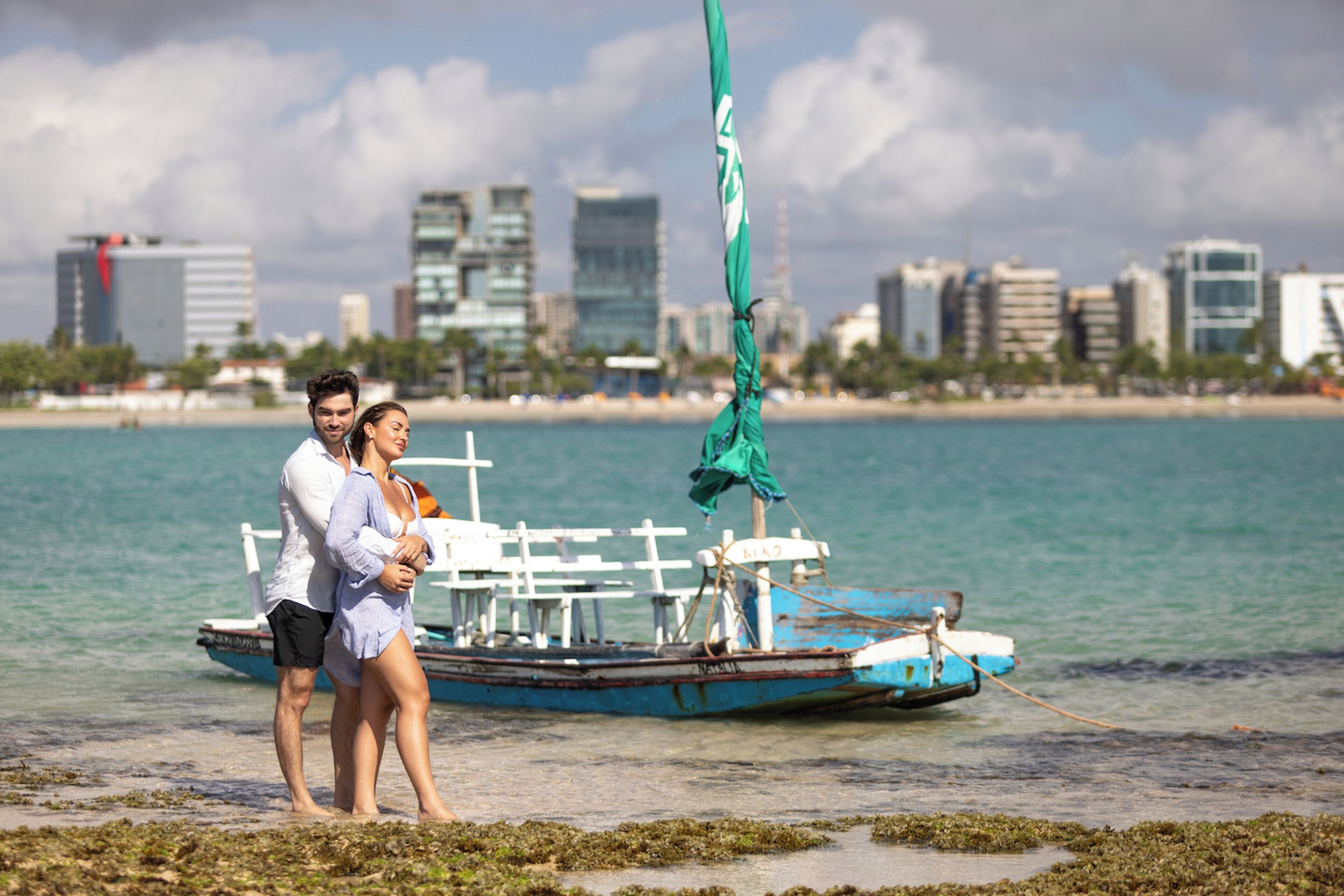 A couple standing near a boat with a city skyline in the background.