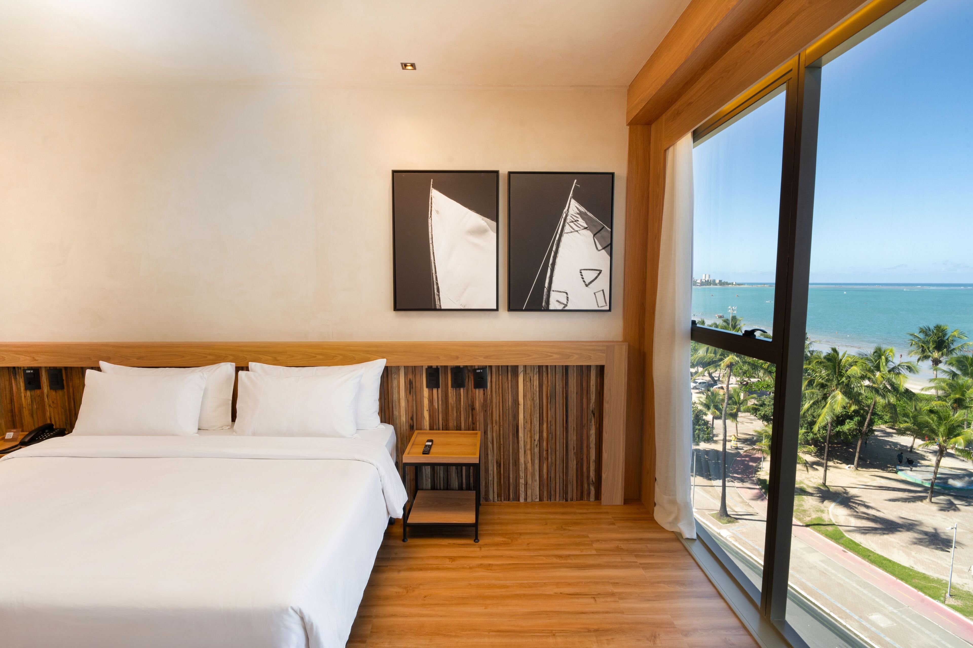 Habitación de hotel con una cama grande, cabecera de madera, mesita de noche, televisor montado en la pared y un ventanal con vistas a la playa y al océano.