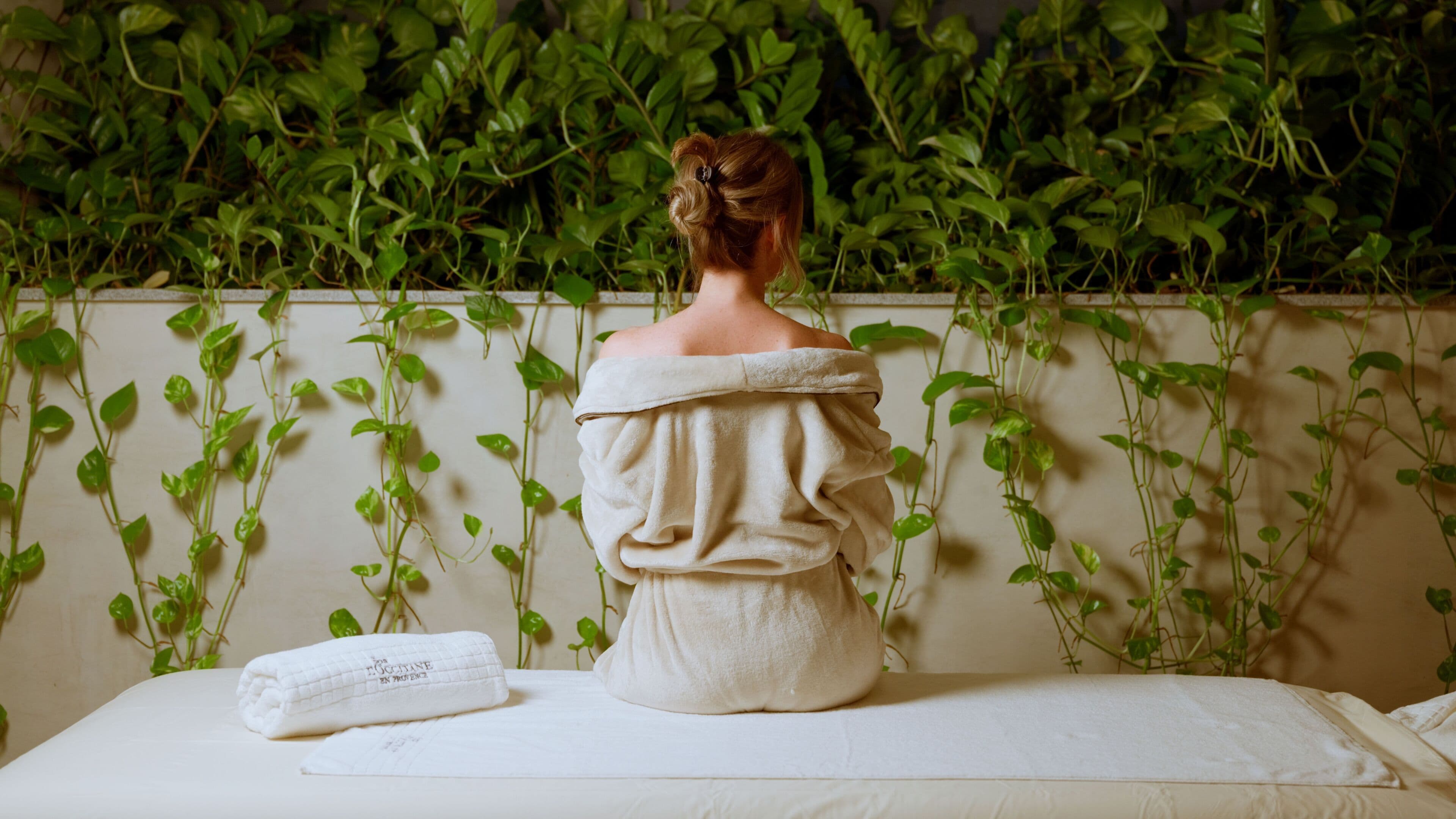 A person seated on a white massage table with a towel beside them, against a wall with green climbing plants.