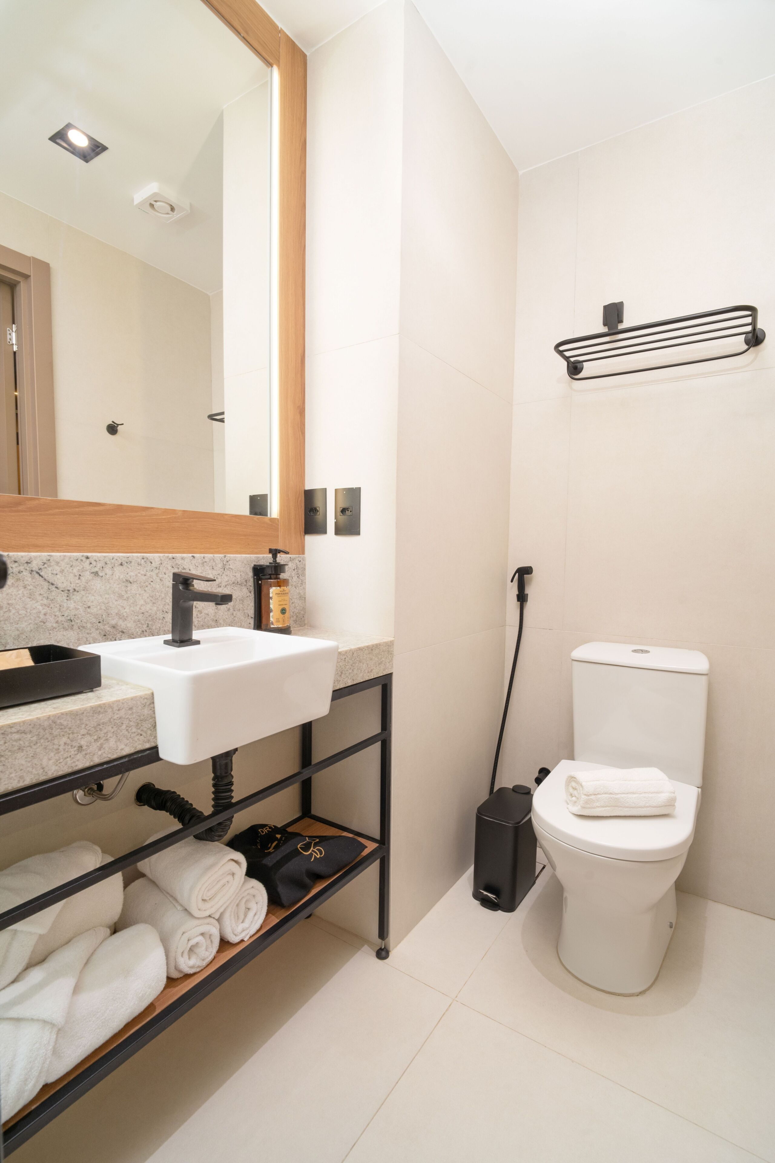 Un baño moderno con un lavamanos blanco, un grifo negro y un inodoro. Hay un estante con toallas y un espejo sobre el lavamanos.