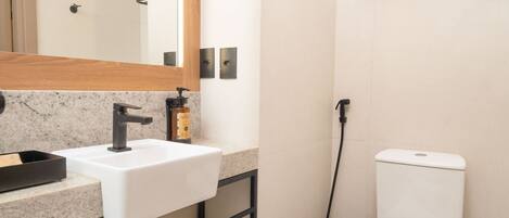 Un baño moderno con un lavamanos blanco, un grifo negro y un inodoro. Hay un estante con toallas y un espejo sobre el lavamanos.