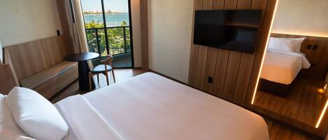 Habitación de hotel con una cama grande, un televisor montado en la pared, una mesita y un balcón con vistas a la ciudad y al mar.