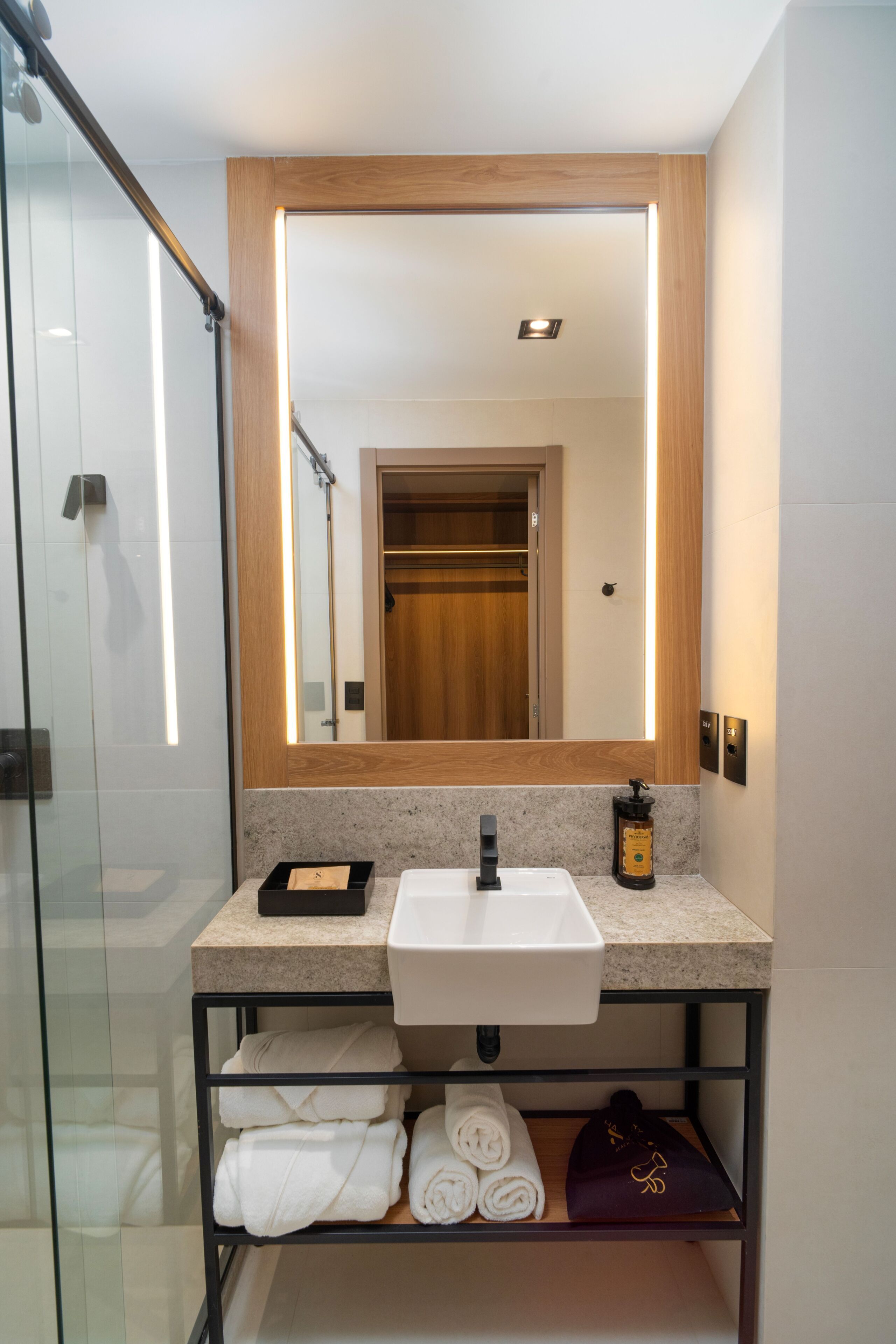 A bathroom with a glass shower enclosure, a white sink, a mirror with a wooden frame, and a shelf with towels.