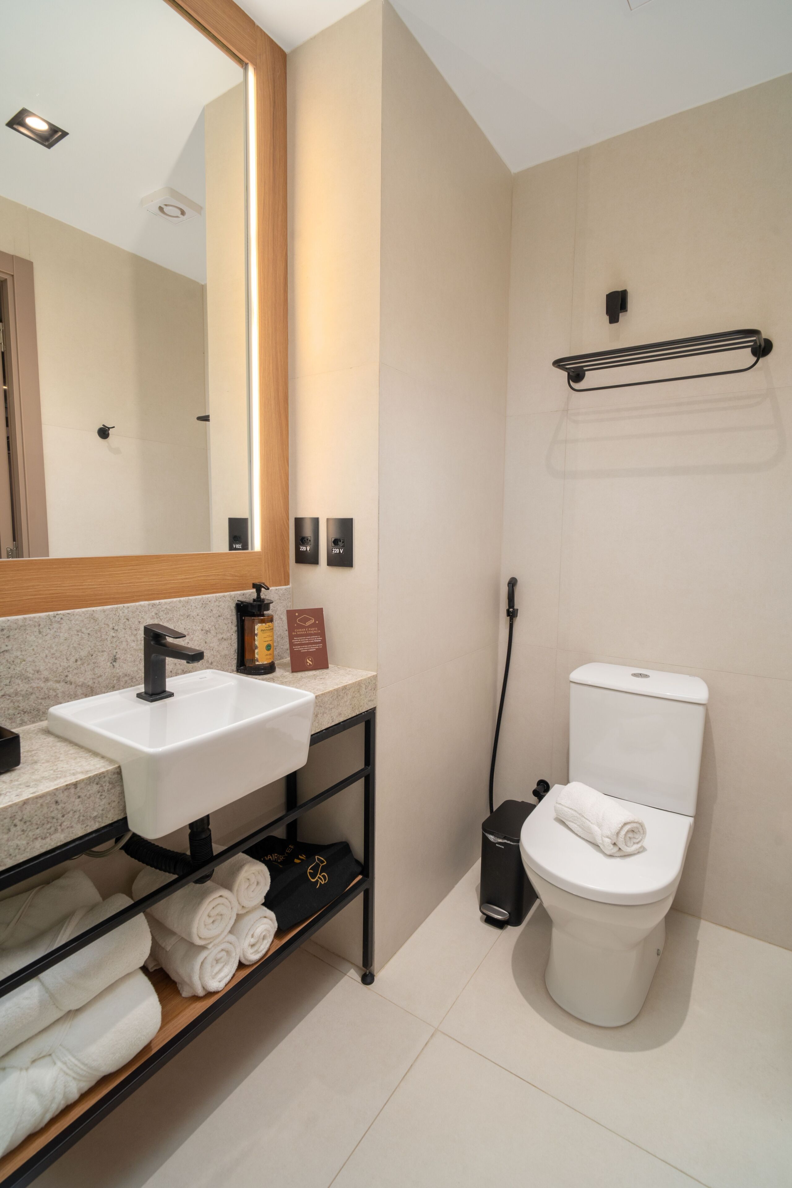 A modern bathroom with a white sink, black faucet, and a toilet. There is a towel rack on the wall and a shelf with folded towels.