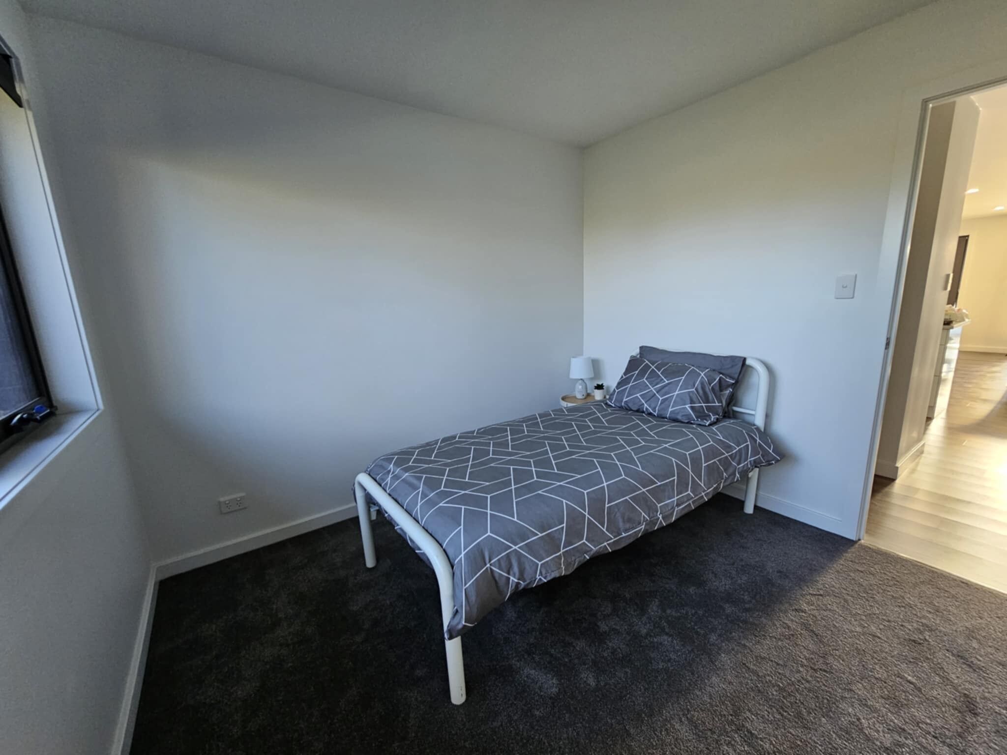 Ein kleines Zimmer mit einem Einzelbett, einer Bettdecke mit grauem geometrischen Muster, einem Nachttisch mit Lampe und einem dunklen Teppichboden.