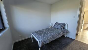 Ein kleines Zimmer mit einem Einzelbett, einer Bettdecke mit grauem geometrischen Muster, einem Nachttisch mit Lampe und einem dunklen Teppichboden.
