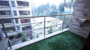 A balcony with a metal railing, a small patch of artificial grass, and a view of a residential area with buildings and palm trees. - 2 Bedroom Apartments (Mumbai)