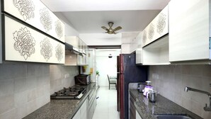 A narrow kitchen with granite countertops, a gas stove, a refrigerator, and decorative wall panels. - 2 Bedroom Apartments (Mumbai)