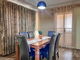 Un comedor con una mesa de madera, seis sillas, un florero con plantas, cortinas estampadas y una ventana con cortinas translúcidas.