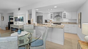 A modern kitchen with white cabinetry, a central island, and a dining area with glass-top table and wicker chairs. - Gulf-Front Grandeur in Longboat Key – Luxurious & Spacious Condo #102 by RVA (Longboat Key)