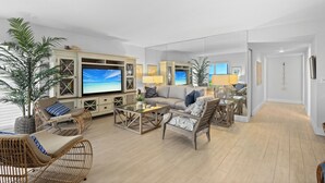 A spacious living room with a large flat-screen TV, comfortable seating, a coffee table, and a large mirror reflecting the room. - Gulf-Front Grandeur in Longboat Key – Luxurious & Spacious Condo #102 by RVA (Longboat Key)