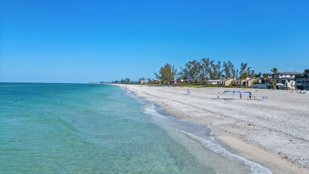 A beach with clear turquoise water, a sandy shore, and a row of buildings with a clear view of the ocean. - Gulf-Front Grandeur in Longboat Key – Luxurious & Spacious Condo #102 by RVA (Longboat Key)