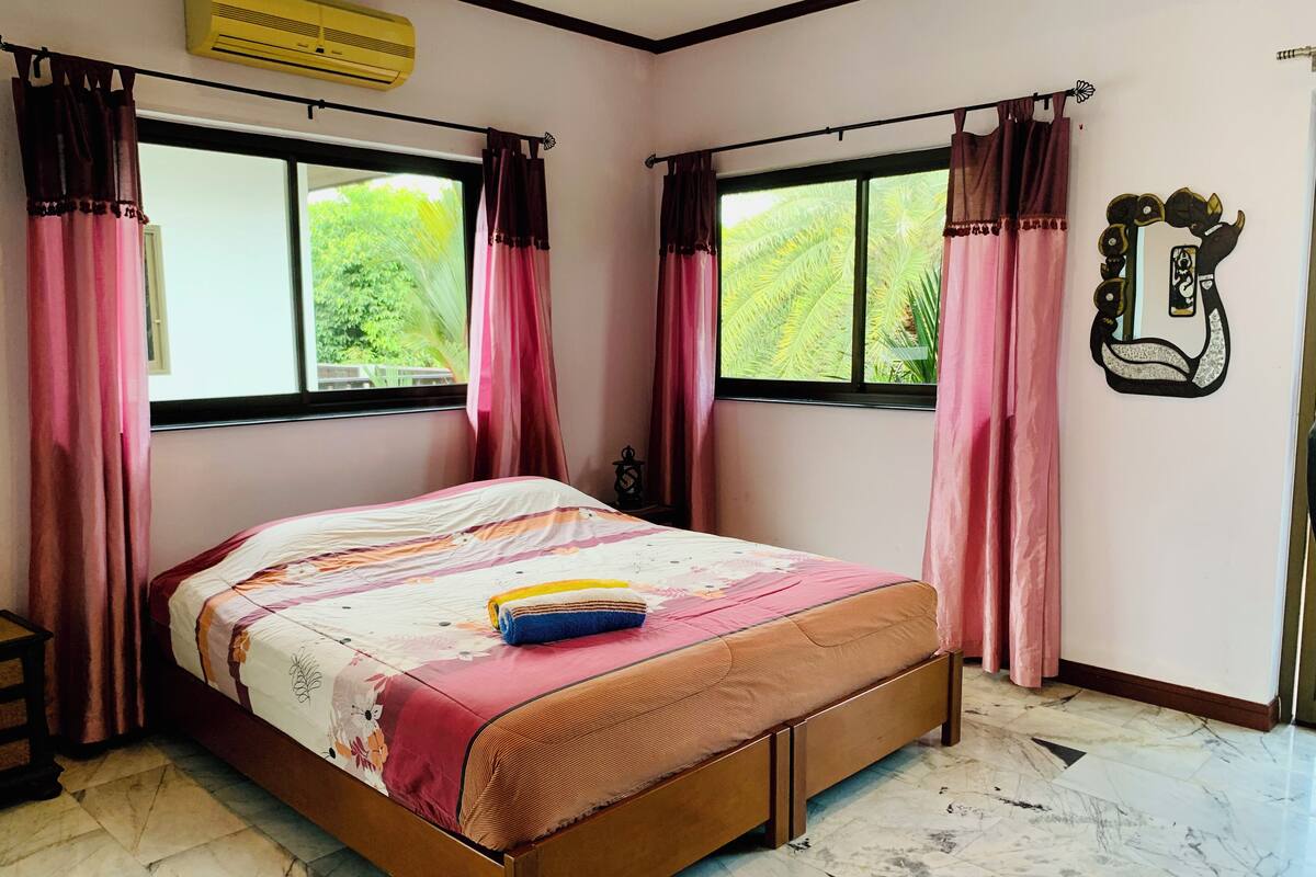 A bedroom with a bed, two windows with curtains, a wall-mounted air conditioner, and a decorative wall piece.