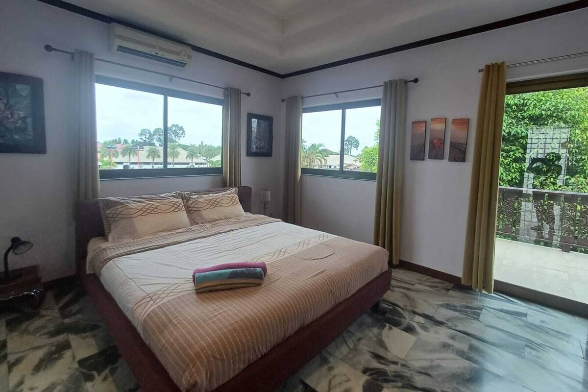 A spacious bedroom with a large bed, two windows with curtains, a ceiling fan, and a view of greenery outside.