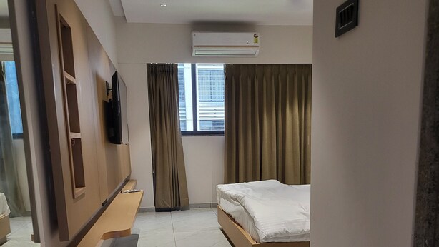 A hotel room with a bed, a TV mounted on the wall, a window with curtains, and an air conditioning unit. - Hotel Shiv Kartik (Surat)