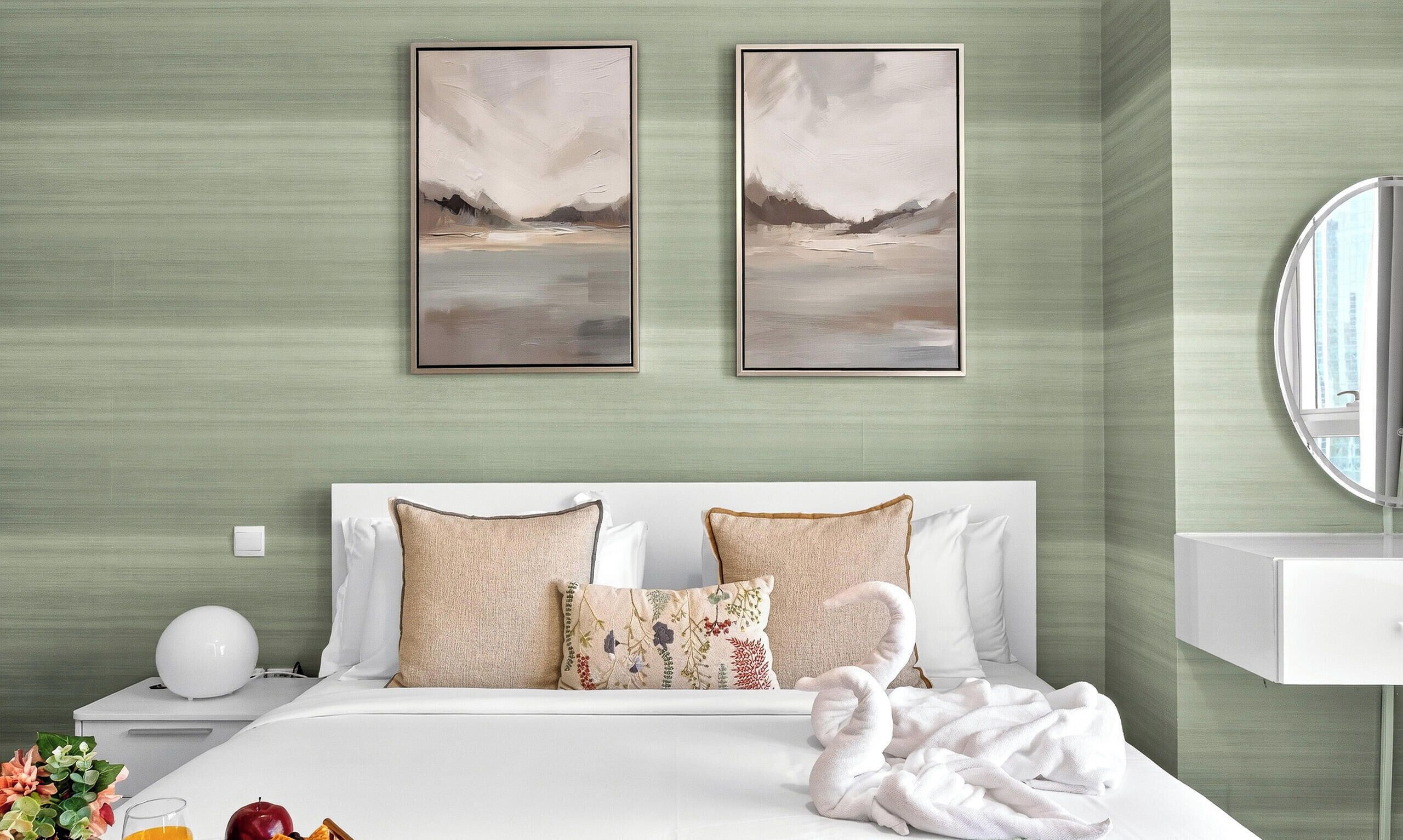 Une chambre d’hôtel avec un lit, deux tableaux accrochés au mur, une table de chevet avec une lampe, un miroir rond et une vue sur un immeuble par la fenêtre.