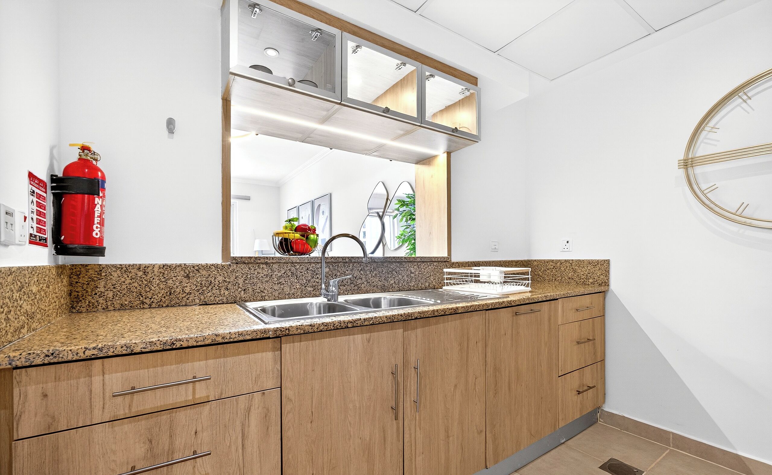 A kitchen with granite countertops, stainless steel sink, wooden cabinets, a fire extinguisher, and a mirror above the sink.