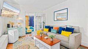 A living room with a sofa, coffee table, and a TV. There is a window with curtains, a radiator, and a plant on the coffee table. - 3BR Charming Beach Cottage | North End OC (Ocean City)