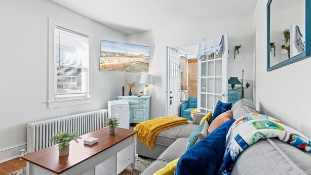 A modern living room with a sofa, a coffee table, a radiator, a window with blinds, a picture on the wall, and a door leading to another room. - 3BR Charming Beach Cottage | North End OC (Ocean City)