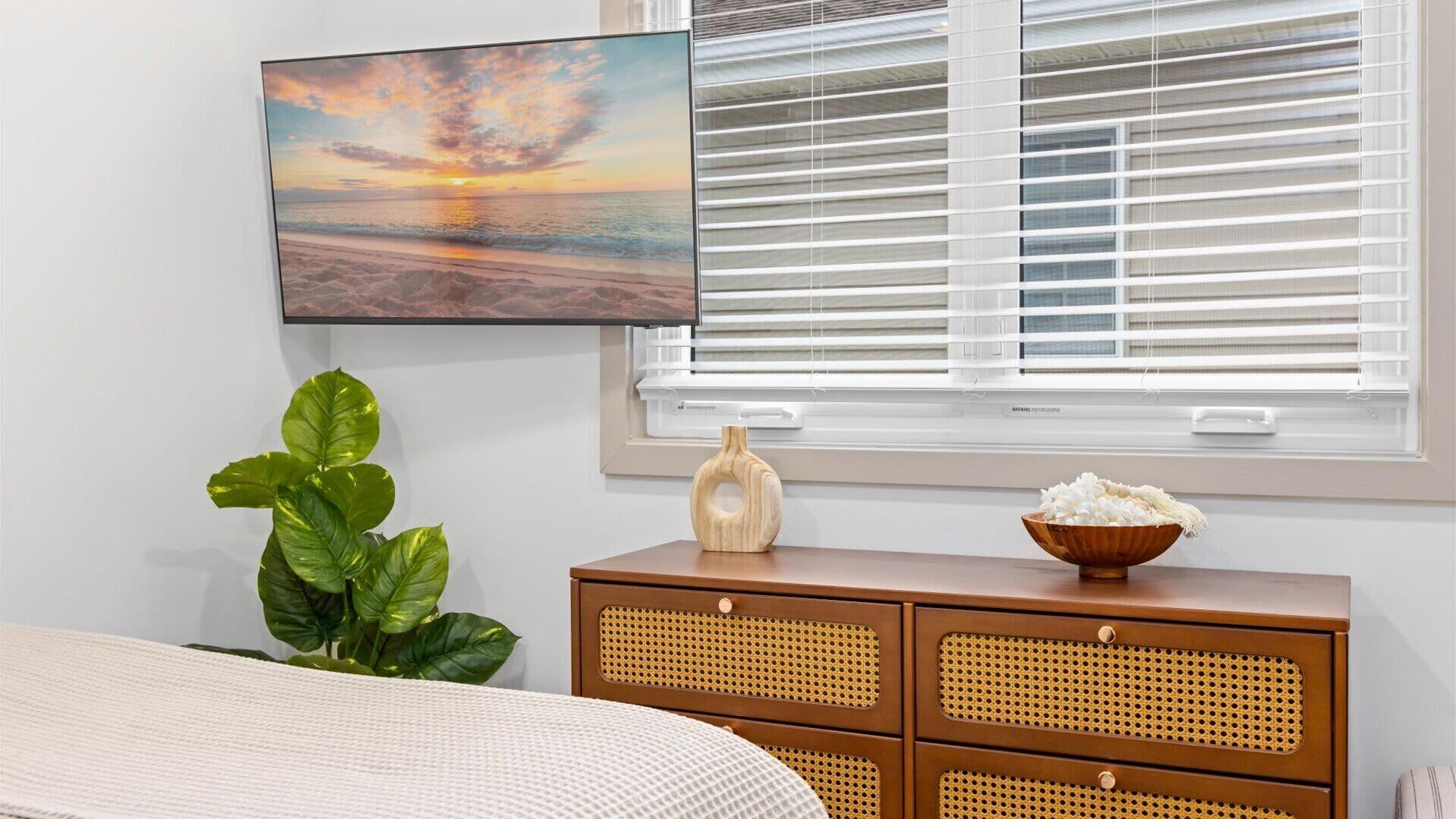 Een slaapkamer met een bed, een grote potplant, een houten dressoir met een schaal rijst, en een schilderij van een zonsondergang.
