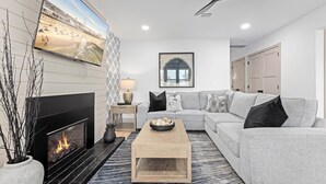 A modern living room with a fireplace, a grey sofa, a coffee table, and a TV displaying a beach scene. - 3BR Modern 2nd Floor Condo | Easy Walk to Beach! (Ocean City)