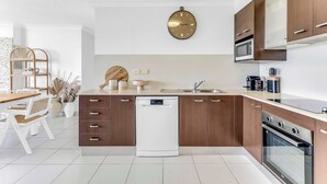 A modern kitchen with wooden cabinets, a dishwasher, a microwave, and a clock on the wall. - Tiki by Khove (Surfers Paradise)