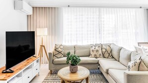 A modern living room with a flat-screen TV, a beige sofa, a round coffee table with a plant, and a large window with sheer curtains. - Tiki by Khove (Surfers Paradise)