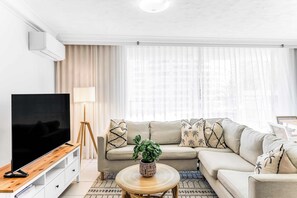 Un salon moderne doté d’un téléviseur à écran plat, d’un canapé beige, d’une table basse ronde agrémentée d’une plante et d’une grande fenêtre ornée de rideaux légers.