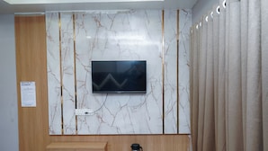 A modern room with a marble-patterned wall, a flat-screen TV mounted above a wooden cabinet, and a glass coffee table with a coffee maker. - Hotel Pleasure (Ujjain)