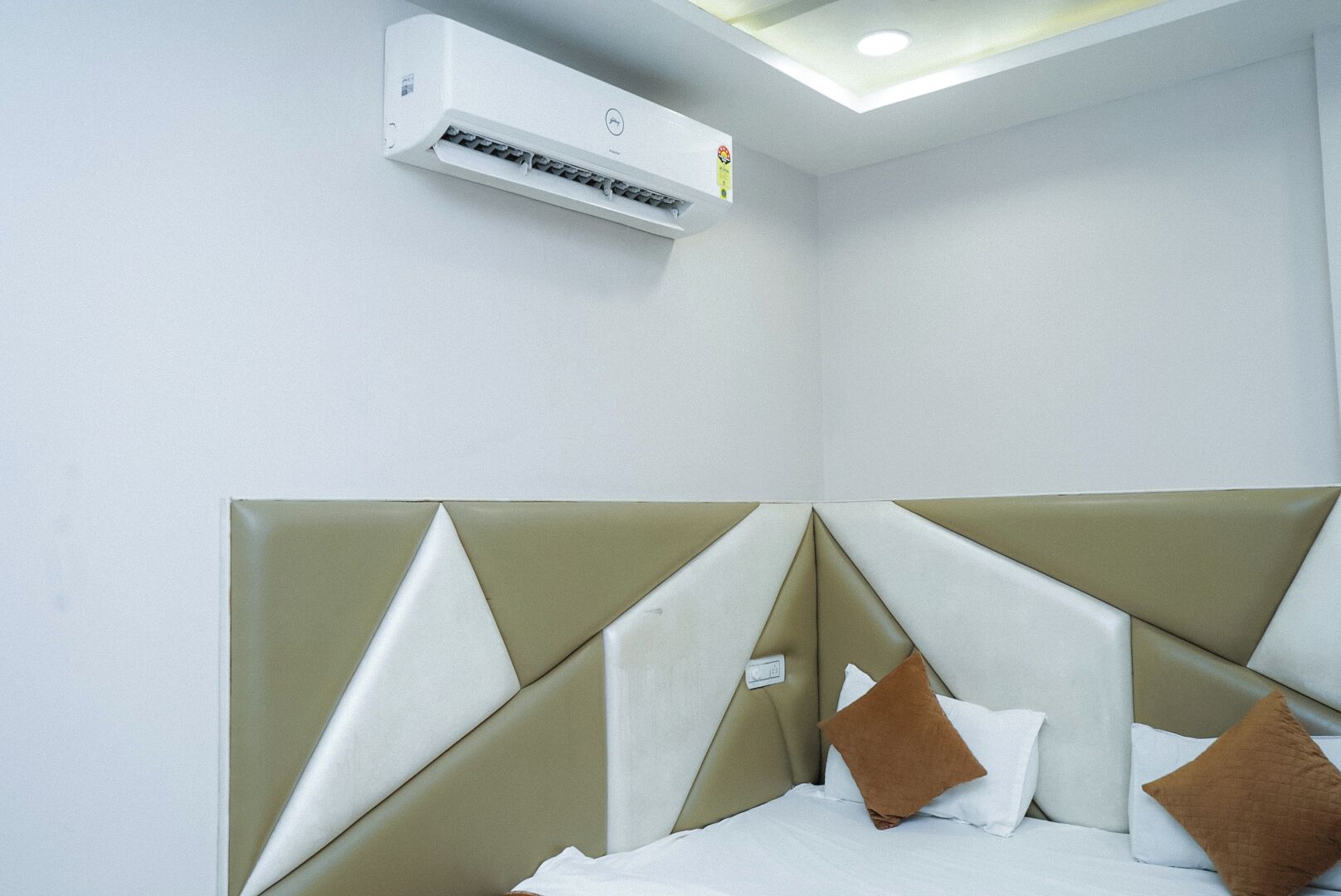 A hotel room with a modern design, featuring a bed with a geometric headboard and two pillows, a white air conditioning unit mounted on the wall, and a ceiling light.