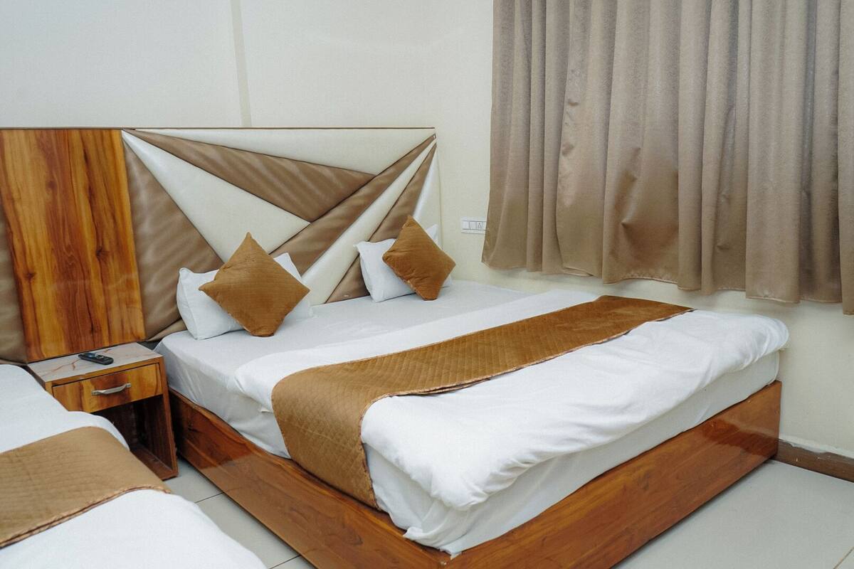 Une chambre d’hôtel avec une tête de lit en bois, un lit avec une literie blanche et des oreillers marron, une table de chevet avec une télécommande et une fenêtre avec des rideaux.