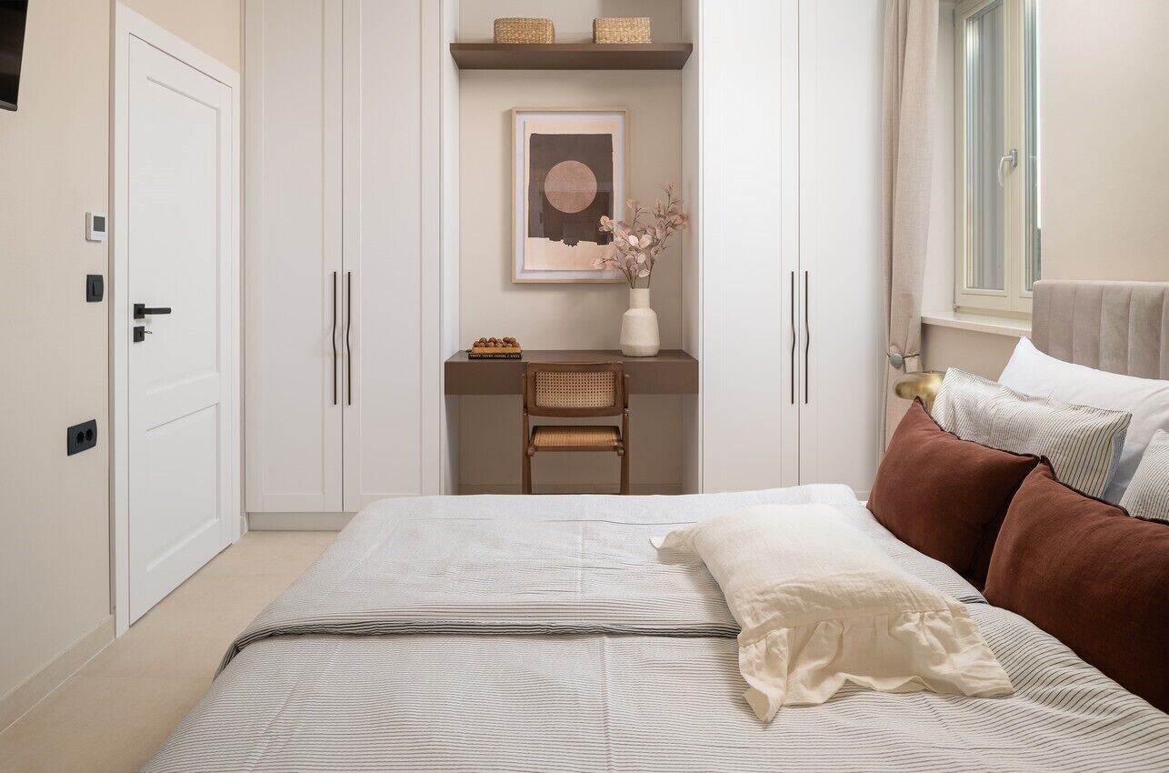 Un dormitorio con cama, mesita de noche, silla, estante con objetos decorativos y ventana con cortinas.