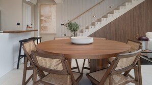 A modern dining area with a round wooden table, six woven chairs, and a central white vase. A staircase with wooden steps and glass railings is visible in the background.
