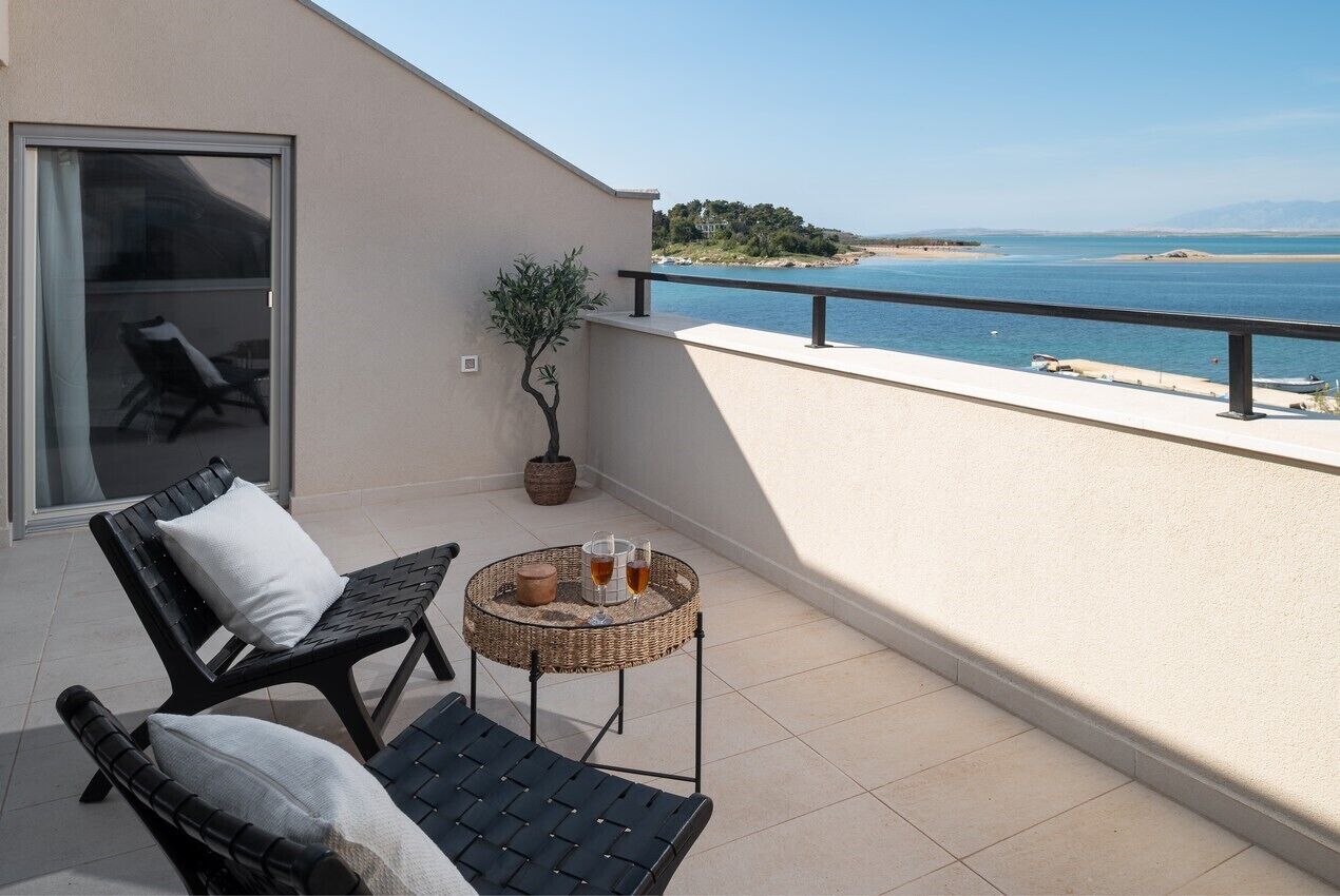 Un balcón con dos sillones de descanso negros, una mesita redonda con bebidas, una planta en maceta y vistas al mar y la playa.