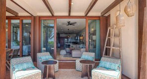 A spacious living area with a large sliding glass door leading to a kitchen and dining area. There are two round wooden tables and four patterned sofas arranged around them.